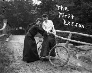 Two women in 1890's style clothing. One is sitting on a bicycle. The other is helping her stay upright. Both are smiling.