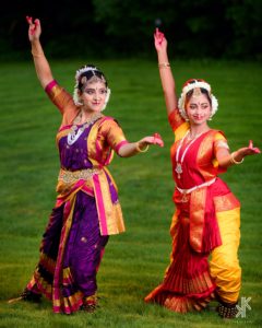 two smiling Kuchipudi dancers pose wearing bright sari in purple, yellow, and red